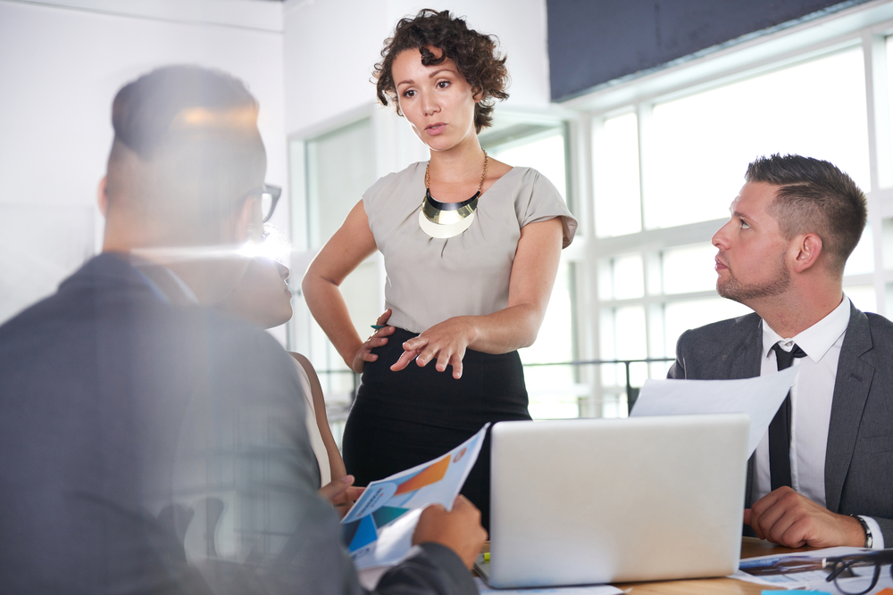 A woman standing, speaking to a few men sitting down at laptops