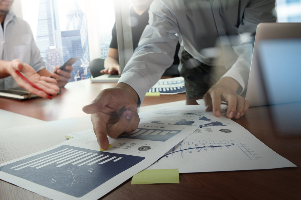 A man pointing at a bar chart that is located on a printed piece of paper located on a desk